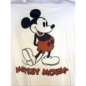 May include: Cream-colored t-shirt featuring a classic Mickey Mouse graphic. The cartoon character is depicted in black, red, and white, with the words "Mickey Mouse" printed in brown below the image. A vintage-style design.