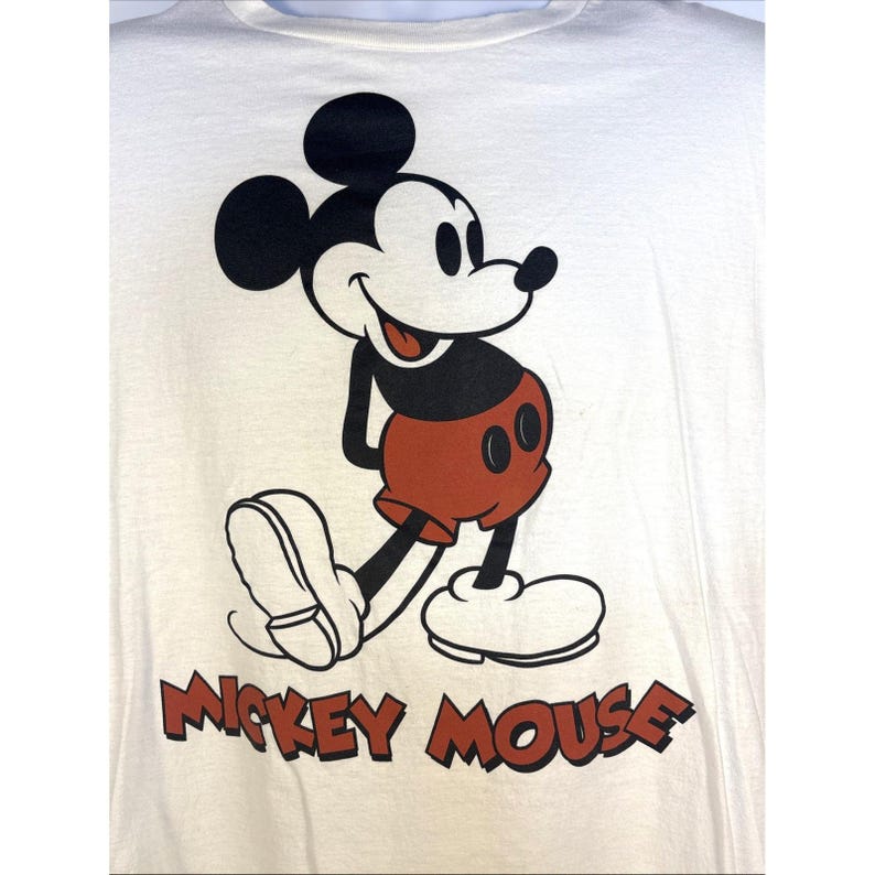 May include: Cream-colored t-shirt featuring a classic Mickey Mouse graphic. The cartoon character is depicted in black, red, and white, with the words "Mickey Mouse" printed in brown below the image. A vintage-style design.