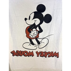 May include: White t-shirt featuring a classic Mickey Mouse graphic. The cartoon character is depicted in black, red, and white, with the words "Mickey Mouse" in red, printed in reverse at the bottom. The design is a vintage style.