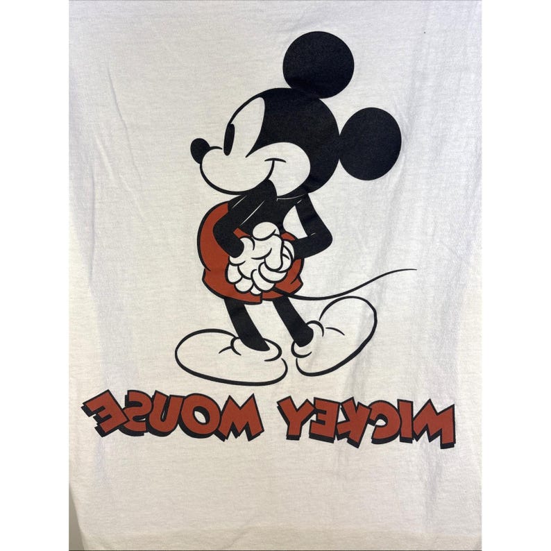 May include: White t-shirt featuring a classic Mickey Mouse graphic. The cartoon character is depicted in black, red, and white, with the words "Mickey Mouse" in red, printed in reverse at the bottom. The design is a vintage style.