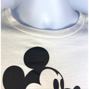 May include: Close-up of a white t-shirt featuring a large black Mickey Mouse graphic. The shirt has a crew neck and is made of a soft, light-colored fabric. The graphic is a bold, simple design.