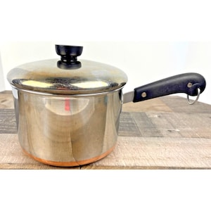 Puede incluir: Un cazo de acero inoxidable con base de color cobre, tapa a juego con pomo negro y mango negro que incluye una pequeña anilla metálica para colgar. El recipiente tiene un acabado pulido y reflectante con un cuerpo cilíndrico.