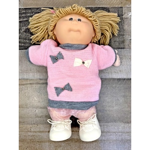 Könnte beinhalten: Eine Puppe mit blondem Wollhaar in Zöpfen, bekleidet mit einem rosa Strickpullover mit grauen Abschlüssen und drei kleinen Schleifen in Grau und Weiß. Die Puppe hat helle Haut, dunkle Augen, eine hellrosa Hose und weiße Schnürschuhe mit Struktur.