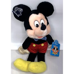 May include: A plush Mickey Mouse toy wearing red shorts with yellow buttons, a blue bow tie, and yellow shoes. The toy has a black nose, large black ears, and a big smile. The toy is holding a small white glove in its right hand. The toy is tagged with "Applause's Disney's Mickey Mouse Pals".