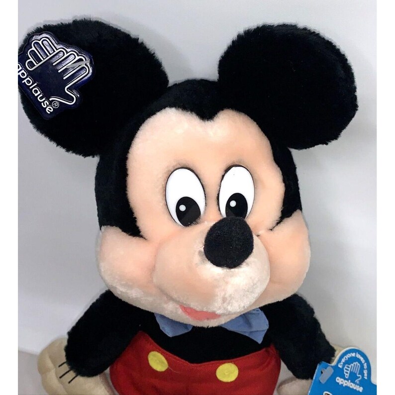 May include: A plush black and white Mickey Mouse toy with a red and yellow shorts, blue bow tie, and a white face. The toy has a tag that says "Applause" on it.