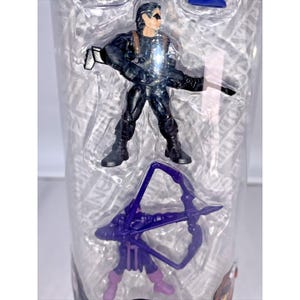 May include: A clear plastic container with a purple bow and arrow, and a black action figure of a superhero with a gun.