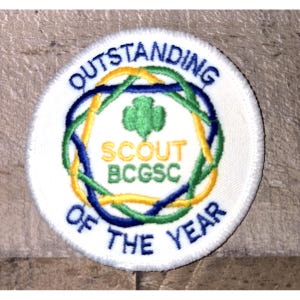 May include: A white, circular patch with the words "OUTSTANDING OF THE YEAR" in blue embroidery. The center features a green shamrock and the text "SCOUT BCGSC" in yellow. Blue and yellow intertwined lines encircle the center.