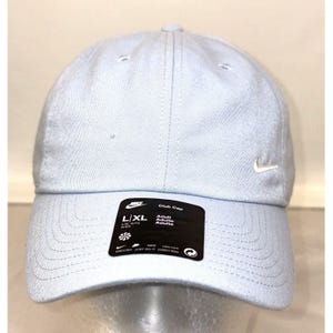 Puede incluir: Una gorra de béisbol azul claro con un logotipo blanco de Nike en forma de swoosh en el lado derecho. La gorra tiene una etiqueta negra en la parte delantera que dice "Club Cap" y "L/XL Adult Adulte Adulto".