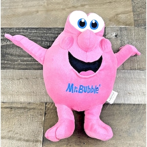 Puede incluir: Peluche rosa del personaje de Mr. Bubble con los brazos extendidos, ojos azules y una gran sonrisa. El personaje tiene una nariz grande y pies. Las palabras "Mr. Bubble" están bordadas en azul.
