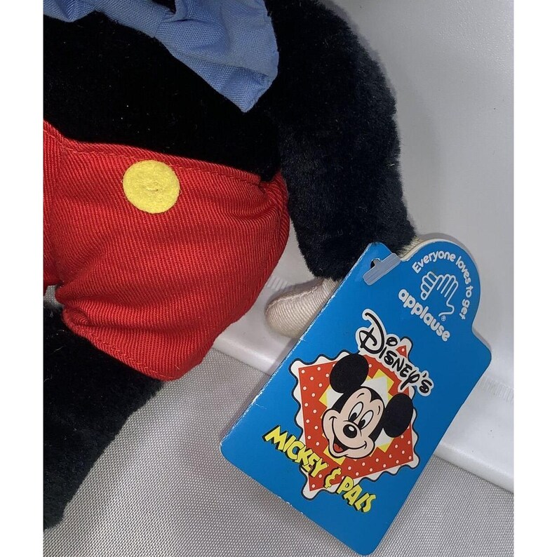 May include: A plush Mickey Mouse toy wearing red shorts with a yellow button and a blue bow tie. The toy is sitting on a white surface with a blue and red tag that says "Disney's Mickey & Pals" and "Everyone loves to get applause".