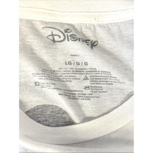 May include: White t-shirt with a gray Disney logo at the top. The shirt's label includes size information, washing instructions, and the text "100% COTTON". The shirt is made in Mexico.