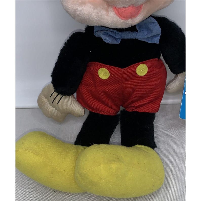 Puede incluir: Un juguete de peluche de Mickey Mouse con pelaje negro, pantalones cortos rojos con botones amarillos y una pajarita azul. El juguete tiene zapatos amarillos y una cara blanca con ojos negros y una boca roja.