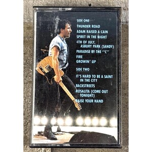 Puede incluir: Una cinta de cassette vintage con una actuación musical en vivo. La portada muestra a un músico con una guitarra y una lista de canciones que incluye "Thunder Road" y "Backstreets". La cinta está en una caja negra.