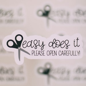 May include: White sticker with black text and a pair of scissors. The text reads "easy does it please open carefully!"