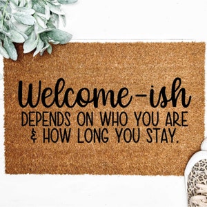 May include: A brown coir doormat with the text "Welcome-ish DEPENDS ON WHO YOU ARE & HOW LONG YOU STAY."