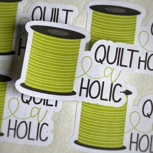 May include: A set of four green and white stickers with a spool of thread and the text "Quilt a Holic".