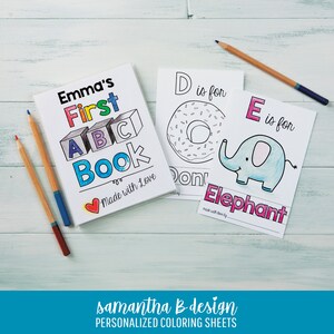 May include: A personalized coloring book titled Emma's First ABC Book with a heart icon, alongside two coloring sheets featuring the letters D for Donut and E for Elephant, accompanied by three colored pencils on a light wooden surface.