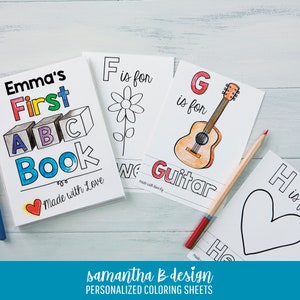 May include: A personalized coloring book titled Emma's First ABC Book with a red heart icon, alongside individual coloring sheets featuring the letters F, G, and H with illustrations of a flower, a guitar, and a heart, plus a colored pencil.