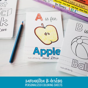 May include: A set of white paper coloring sheets featuring alphabet letters and illustrations. One sheet shows a colored apple with the text A is for Apple and made with love by aunt Donna. A blue pencil lies next to the sheets on a white surface.