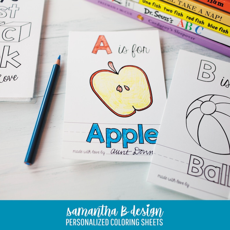 May include: A set of white paper coloring sheets featuring alphabet letters and illustrations. One sheet shows a colored apple with the text A is for Apple and made with love by aunt Donna. A blue pencil lies next to the sheets on a white surface.