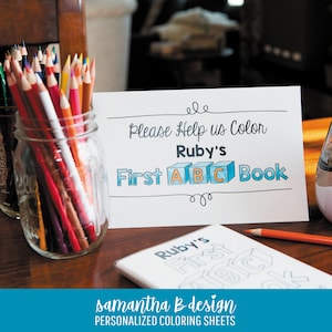May include: A white sign with black text that reads "Please Help us Color Ruby's First ABC Book" with the letters A, B, and C inside blue cubes. A glass jar filled with multicolored pencils sits next to a coloring book with the same title.