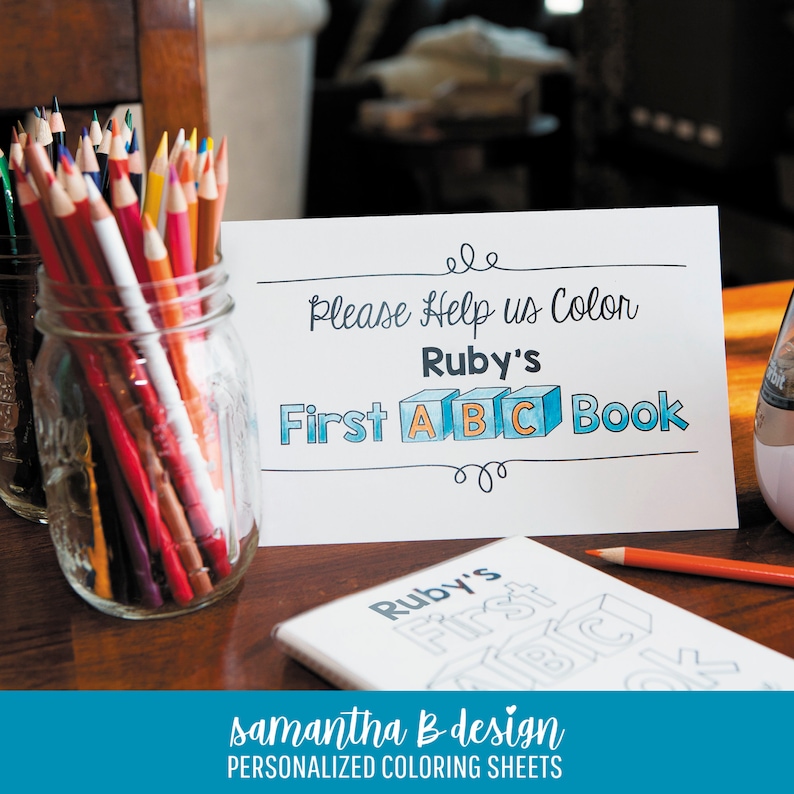 May include: A white sign with black text that reads "Please Help us Color Ruby's First ABC Book" with the letters A, B, and C inside blue cubes. A glass jar filled with multicolored pencils sits next to a coloring book with the same title.