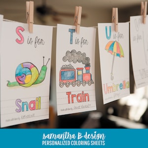 May include: A series of white paper coloring sheets hanging from a string with wooden clothespins. Each sheet features a letter of the alphabet, a hand-colored illustration of a snail, train, or umbrella, and the corresponding word in colorful letters.