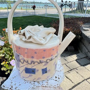 May include: A ceramic teapot with a high arched handle and a lid featuring a sculpted white rose. The body is cream-colored with a pink band at the top decorated with purple dots, and the lower section features blue rectangles and purple squiggles.