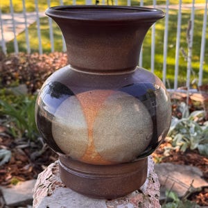 May include: A ceramic vase with a matte dark brown finish on the neck and base. The rounded body features a glossy glaze with large, overlapping circular patterns in shades of muted orange, beige, and deep black, creating a distinct contrast.