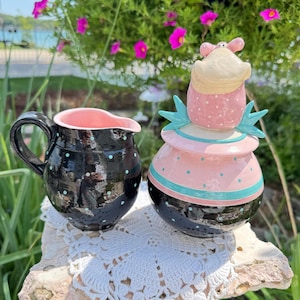 May include: A black ceramic creamer with light blue polka dots and a pink interior, paired with a matching lidded sugar bowl featuring a pink top, light blue accents, and a small sculpted figure on the lid, both resting on a white lace doily.