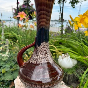 May include: A tall ceramic pitcher vase with a glossy finish featuring a gradient of deep brown, black, and cream colors. The vessel has a rounded base, a slender neck, and a curved handle on one side, showcasing a textured drip glaze pattern.
