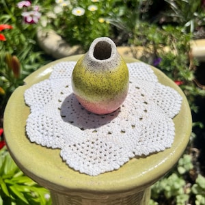 May include: A small, rounded ceramic vase featuring a speckled texture with a color gradient transitioning from a pale pink base to a vibrant yellow midsection and a light, speckled top opening. The vase sits centered on a white crocheted doily.