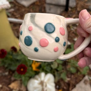 May include: A ceramic mug with a cream-colored base featuring a subtle gray marbled swirl pattern. The exterior is decorated with raised, smooth, circular dots in alternating shades of teal and soft pink, complete with a curved handle.