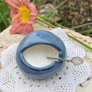 May include: A round, blue ceramic bowl with a textured, speckled glaze and a wide, open top. Inside the bowl is a white, powdery substance. A silver-toned metal spoon with a twisted handle and a small, pale pink stone accent rests inside.