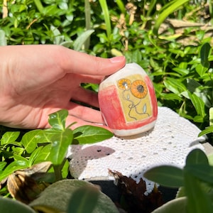 May include: A small, rounded ceramic vessel with a white base, featuring a rectangular panel on the front. The panel contains a yellow background with two hand-drawn orange flowers on thin, dark stems and a single small green leaf.