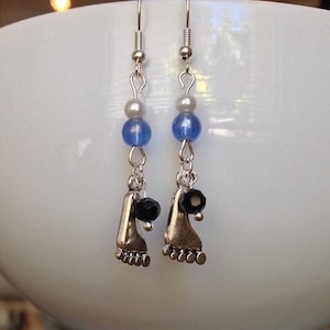 May include: A pair of dangle earrings featuring silver-toned foot charms, blue and gray beads, and black faceted beads. The earrings have silver-colored fish hook ear wires.