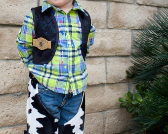 May include: A young boy wearing a green and blue plaid shirt with a black vest and a pair of black and white cow print chaps. He is smiling and looking to the side.
