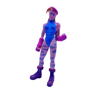 May include: A plastic action figure of a character with blonde braided hair wearing a pink beret, a blue leotard, and blue camouflage leggings. The figure has pink wrist guards and dark purple combat boots, standing in a forward-facing pose.