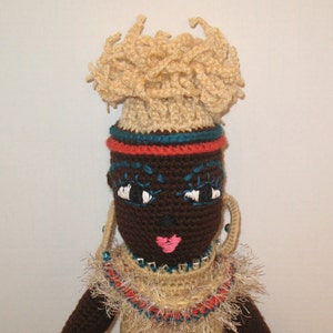 Puede incluir: Muñeca de crochet con piel marrón, cabello negro y un collar de cuentas de colores. La muñeca lleva un vestido de crochet marrón con ribete rojo y azul. La muñeca tiene ojos grandes y negros con pupilas blancas y labios rosados.