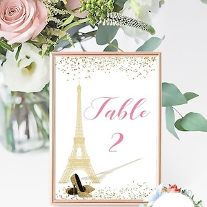May include: A gold glitter table number card with a pink background. The card features a gold silhouette of the Eiffel Tower and the text "Table 2". Two black high heels are at the bottom of the card.