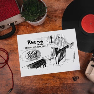 May include: A monochrome sketch of King Tut's music venue, labelled "Best UK Live Venue - Radio 1". The image is on a wooden surface, alongside a vinyl record, a small plant, and a red and black cable.