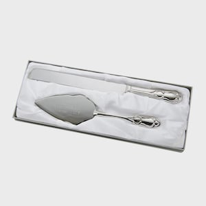 May include: A silver cake knife and server set in a white box. The server is engraved with text. The handles have a decorative design. The set is presented on a white fabric lining.