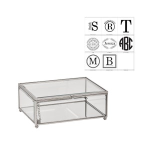 May include: A clear glass box with a silver metal frame and a hinged lid. The box has a small chain attached to the lid. The box is sitting on a white background. There are several different monogram designs in the background.
