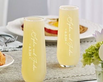 May include: Two clear champagne flutes filled with a yellow beverage, likely a mimosa or similar cocktail. Each glass has the inscription "Sophia and Jordan ~ 6.3.24" in a delicate script. The glasses sit on a table setting with a plate of food.