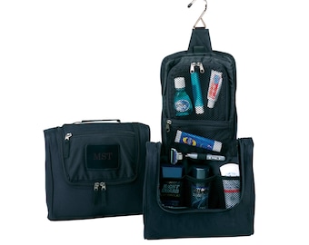 May include: Black travel toiletry bag with a hanging hook, zippered compartments, and a mesh pocket. The bag is open and has various toiletries inside, including a toothbrush, toothpaste, and a bottle of mouthwash. The bag has the initials "MST" embroidered on the front.