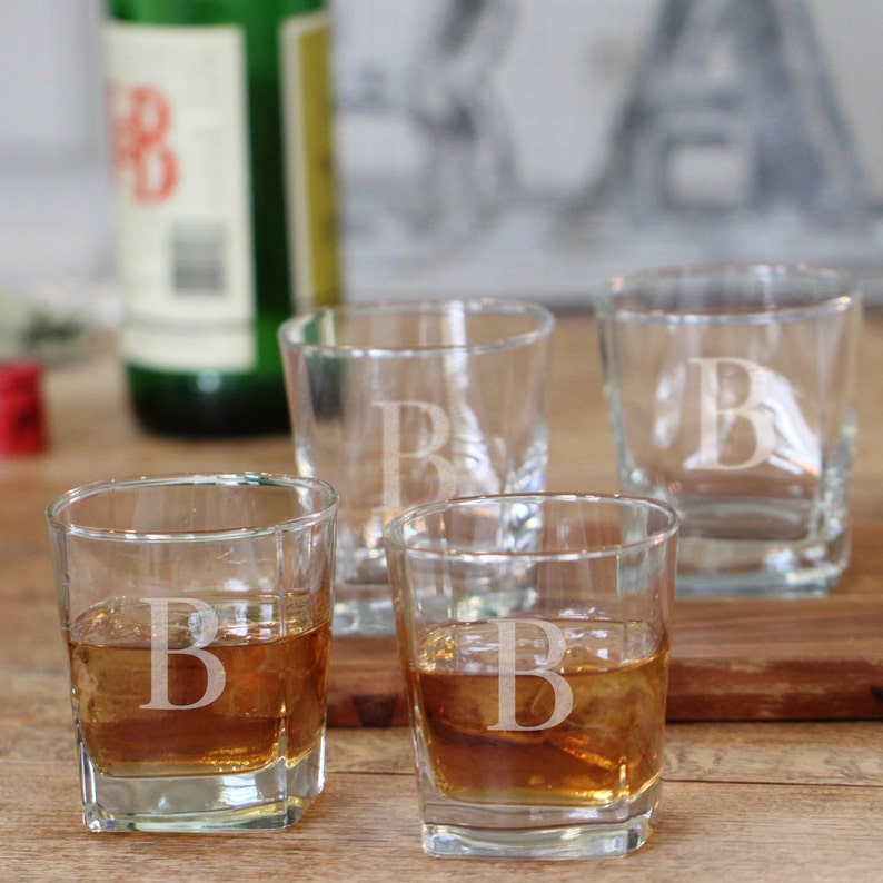 May include: Four clear square-shaped shot glasses with the letter "B" etched on them. Two glasses are filled with a brown liquid, likely a spirit, and ice cubes. The glasses are arranged on a wooden surface, with a green bottle in the background.