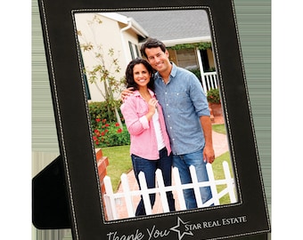 May include: Black leather picture frame with a white stitched border. The frame has a white mat and a photo of a couple standing in front of a house. The frame has the text "Thank You" and "STAR REAL ESTATE" printed on the bottom.