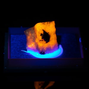 May include: A glowing, rough-textured rock specimen with a dark inclusion, illuminated by a vibrant orange light. The rock sits within a rectangular frame, with a blue glowing base and a dark background.