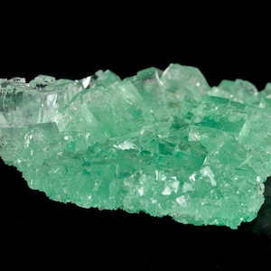 Puede incluir: Un grupo de cristales de fluorita verde claro translúcidos sobre un fondo negro. Los cristales tienen una estructura cúbica y reflejan la luz, creando un efecto brillante. La forma general es irregular y natural.