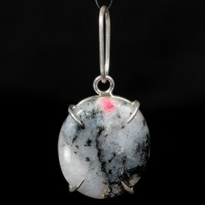 May include: A round pendant with a silver bail and setting. The stone is primarily white with black and grey veining and a small pink spot. The pendant is suspended against a black background.